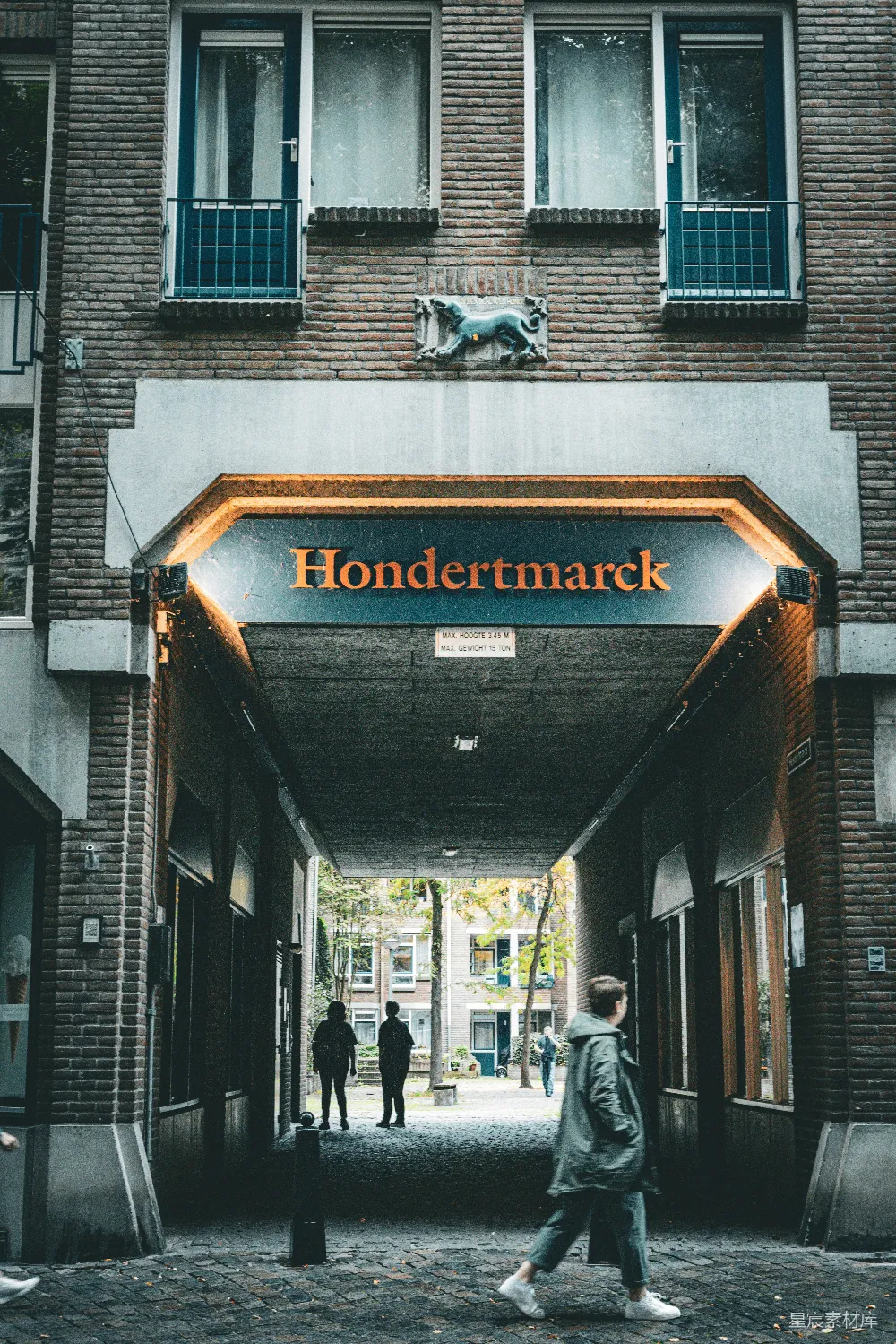 图片展示了一条充满荷兰特色的街道，Hondertmarck的拱门下，几位行人悠然自得地行走，砖砌建筑与人行道的结合，营造出一种宁静而温馨的氛围。背景中的树木和远处的建筑增添了几分自然与现代的融合。