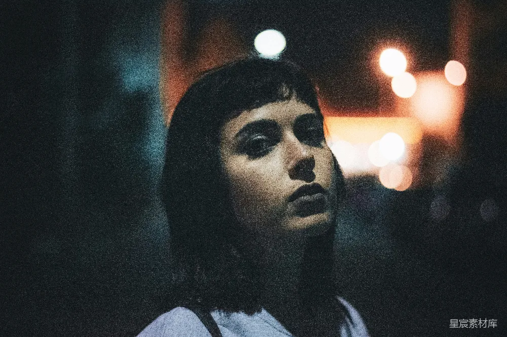 这张图片展示了一个人在夜晚的街道上凝视着前方，背景中有模糊的灯光。人物的侧脸显得有些忧郁，给人一种深思和孤独的感觉。整体色调偏冷，光线昏暗，营造出一种静谧而神秘的氛围。适合用作情感表达或都市生活主题的素材。
