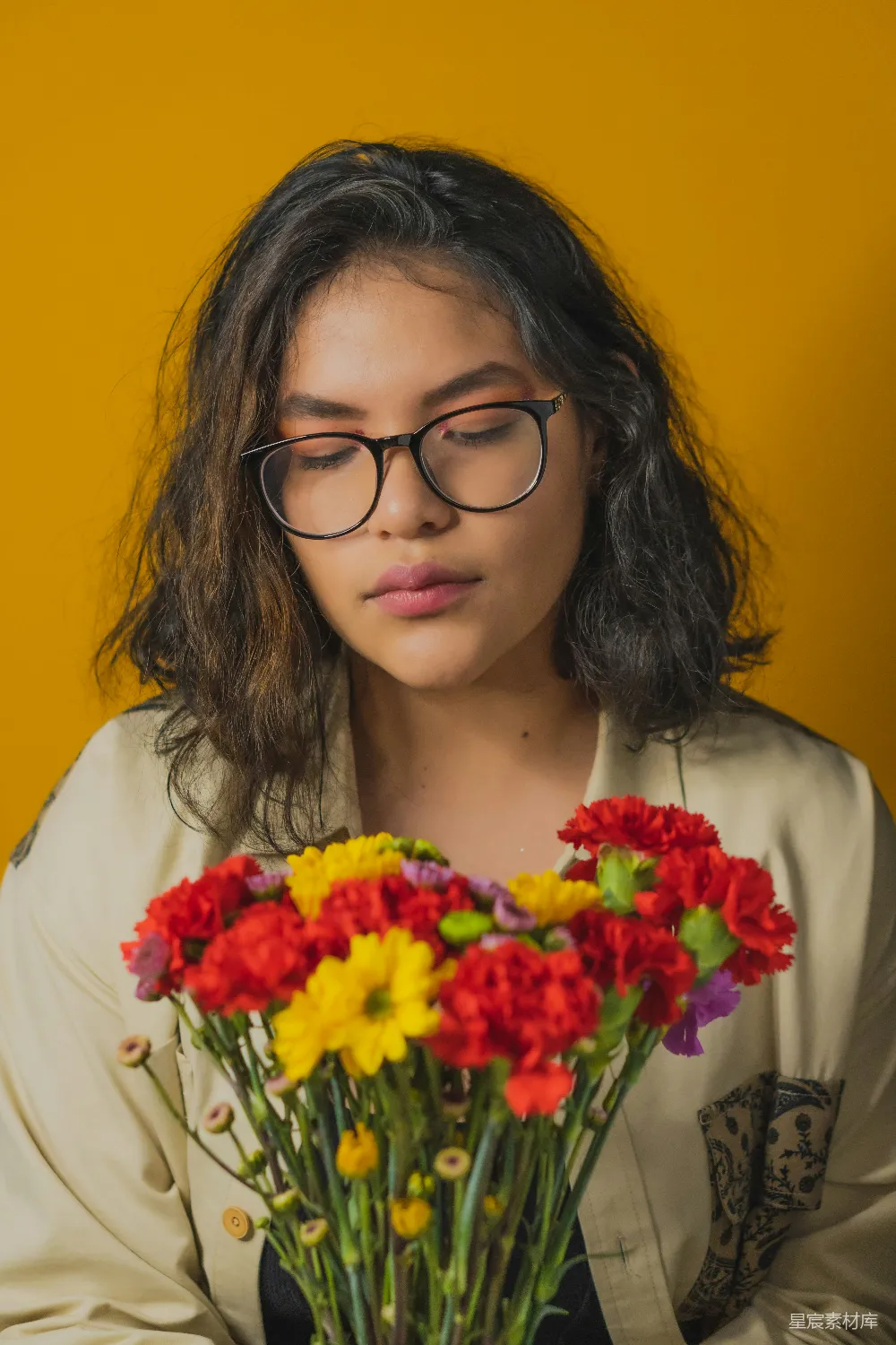 图片中一位戴眼镜的女性，头低下，眼神沉思，手中捧着一束色彩鲜艳的花朵。背景为鲜艳的黄色，整体色调温暖而和谐。适合用于情感表达、艺术创作或个人写真。