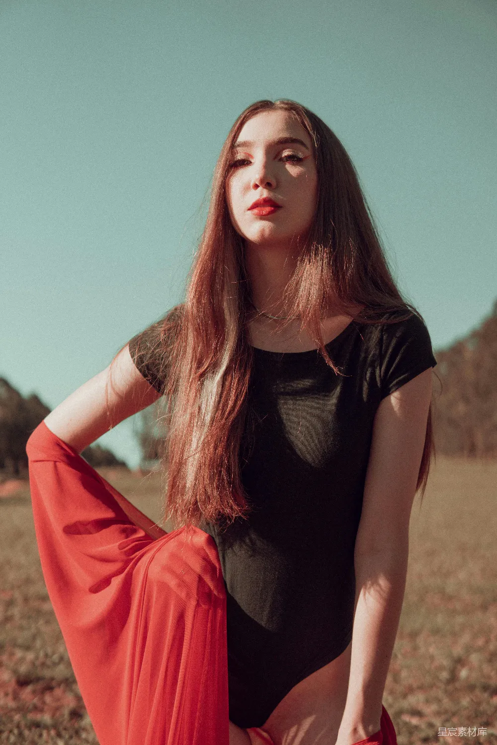 这张图片展示了一位身穿红裙的女孩在阳光下的场景。她站在一片开阔的田野中，背景是蓝天和远处模糊的树木。女孩身着黑色紧身上衣，搭配红色长裙，整体造型时尚而充满活力。阳光洒在她的身上，营造出一种温暖而宁静的氛围。这种场景非常适合用于时尚写真、户外
