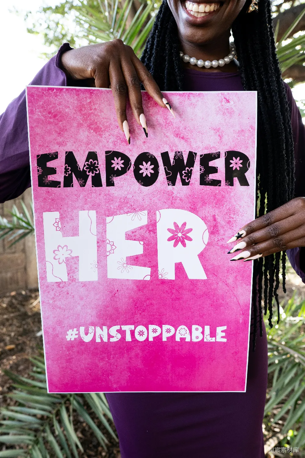图片展示了一位女性手持一张粉色的海报，上面写着“EMPOWER HER #UNSTOPPABLE”，并点缀着小花图案。背景是户外的植物，营造出一种自然和积极向上的氛围。这张图片适合用于宣传女性权益、鼓励女性自我提升和表达对女性力量的支持。