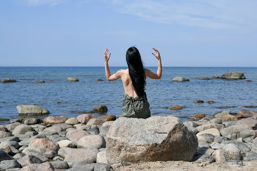 一位女性背对镜头，坐在海边的岩石上，双手轻举，似乎在进行冥想。背景是广阔的海洋和散布的岩石，天空清澈，给人一种宁静与平和的感觉。这幅画面适合用于表现自然、冥想、平静的主题，也适合用作旅行、户外生活或心理健康相关的场景。