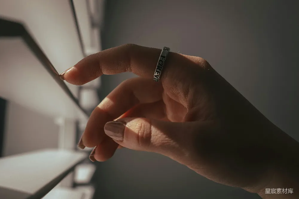 这张图片展示了一只手，手指上戴着一枚刻有“FOREVER”的戒指，背景是柔和的阳光和模糊的室内环境。阳光洒在手上，营造出一种温暖而浪漫的氛围，适合用作时尚生活或情感表达的背景素材。