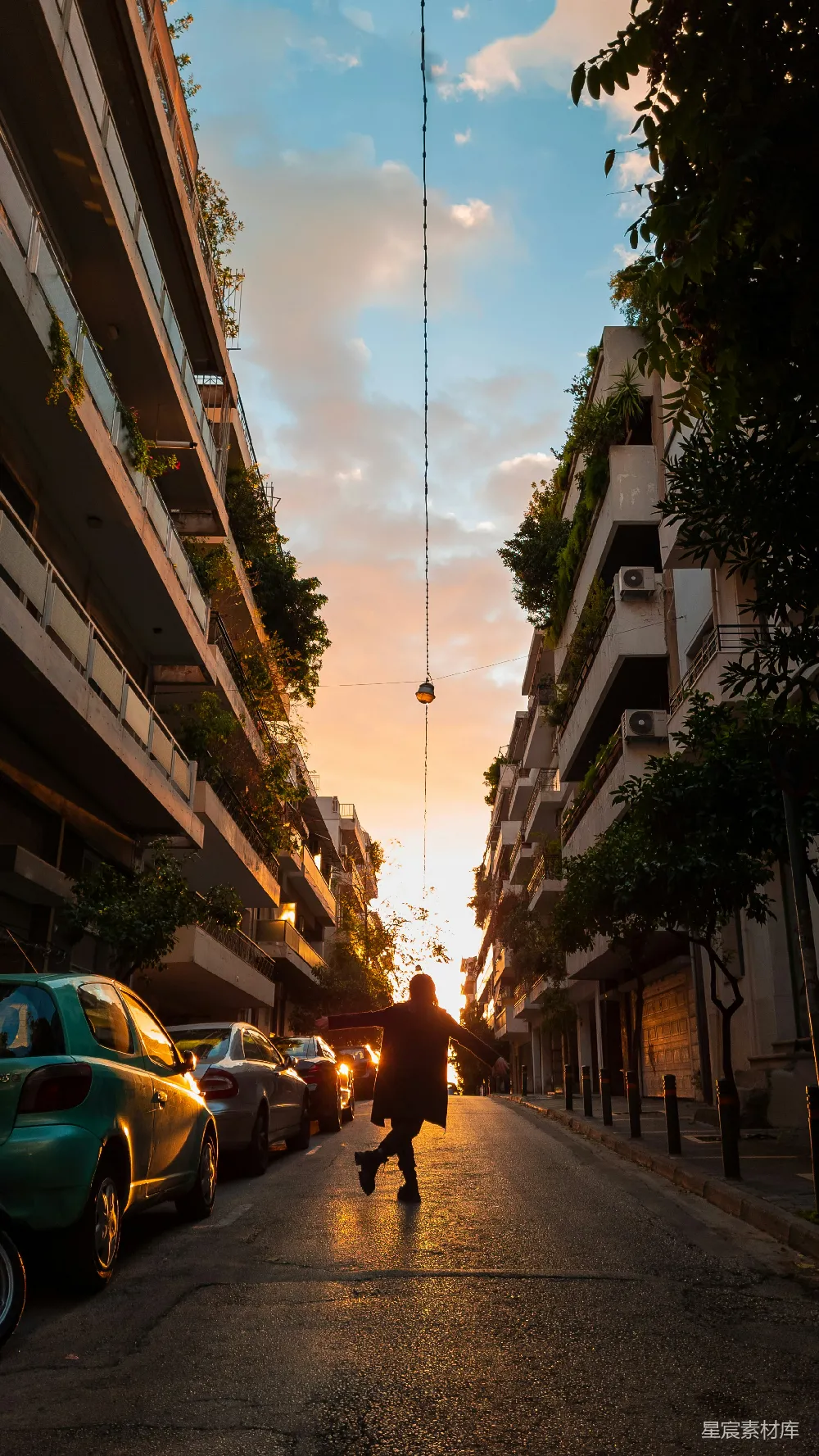 图片展示了一位在城市小巷中奔跑的人，夕阳的余晖洒在街道上，树木和建筑物被染上了温暖的橙色。街道两旁停满了汽车，远处的天空中有几朵云彩，整个场景充满了自由和宁静的氛围。