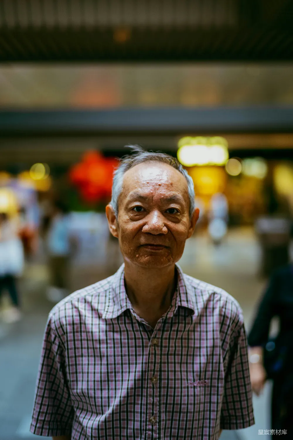 这张图片展示了一位穿着格子衬衫的老人，他站在一个热闹的城市街道上，背景模糊，突显了老人的面部表情和服装细节。光线柔和，营造出一种温馨而略带怀旧的氛围。适合用于城市生活、人物肖像或老年人主题的素材。