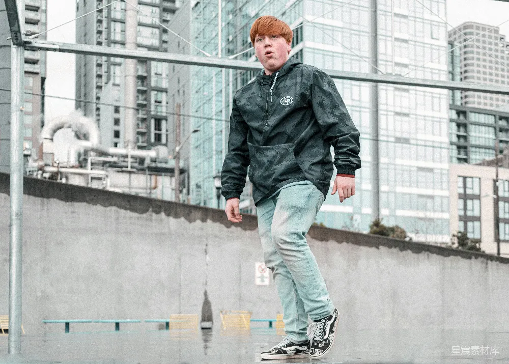 一位年轻男子在城市的屋顶平台上滑板，背景是现代的城市建筑。他穿着迷彩连帽衫和浅色牛仔裤，显得轻松自在。这幅画面捕捉了年轻人在城市中的自由与活力，适合用于展现城市生活、运动休闲或时尚风格的素材。