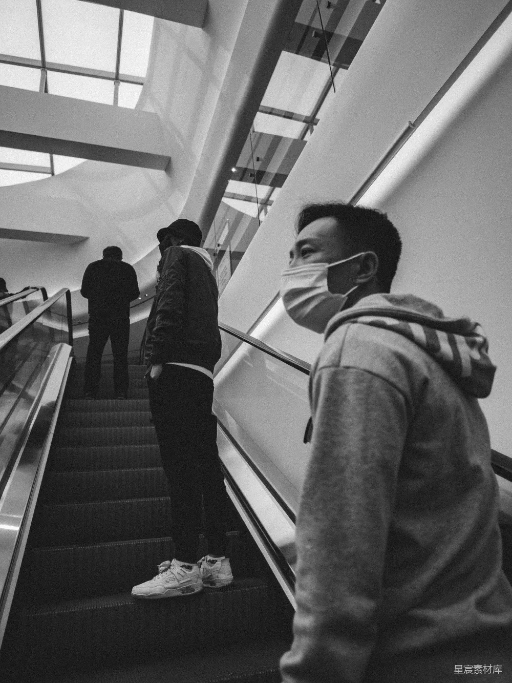 这张黑白照片捕捉了城市中一个普通的瞬间——人们在电梯上匆匆走过。照片的视角从下方仰视，带有一种独特的视角和深度。黑白的色调增加了画面的质感和怀旧感，背景中的高挑建筑和楼梯设计透露出现代都市的气息。这种类型的图片适合用于展现都市生活的多样性和