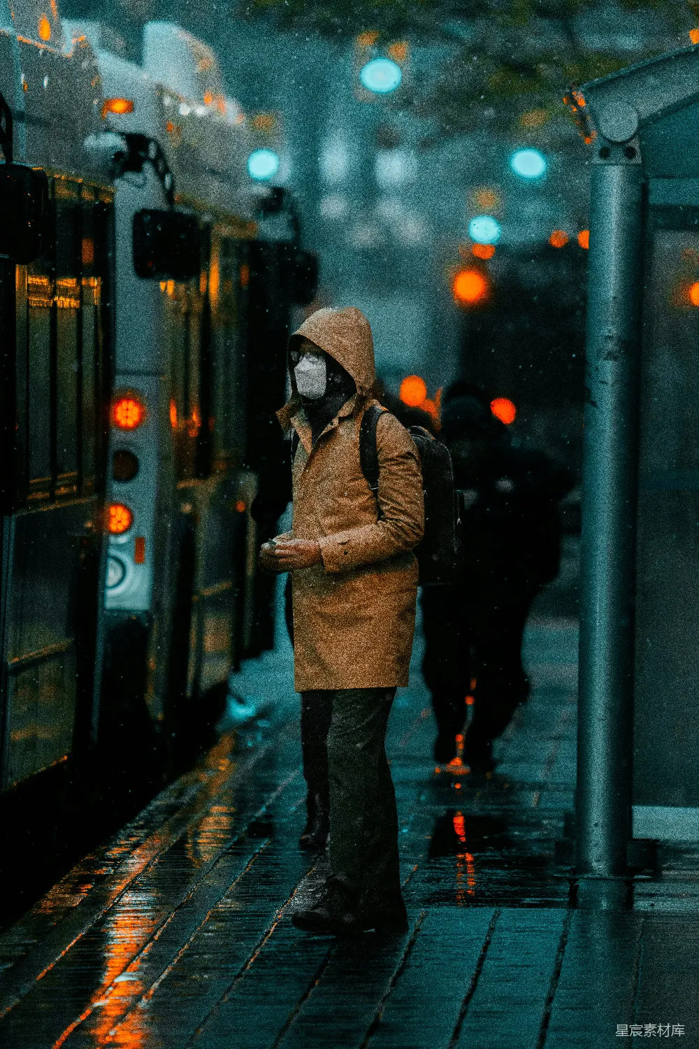 这是一张在雨夜拍摄的街头照片，照片中一位穿着厚实衣物的人站在公交车站台，周围是模糊的灯光和雨滴，营造出一种孤独和宁静的氛围。画面色调偏冷，背景的灯光在雨夜中显得格外温暖，体现了城市夜景的独特美感。