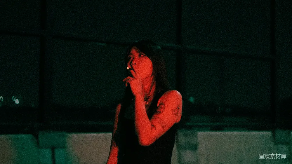 这张图片展示了一个在夜晚独自吸烟的女性，她站在城市的某个角落，背景是昏暗的灯光和模糊的建筑轮廓。她的姿态显得有些孤独和深思，手上的纹身和烟雾的红色调为画面增添了一丝神秘感。这幅图像适合用作情绪深沉、孤独或城市夜景的背景素材。