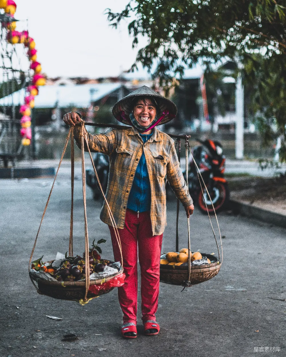 这张图片展示了一位身穿传统服饰的越南街头小贩，她肩上扛着两个装满水果的篮子，面带微笑，显得非常亲切。背景中可以看到节日装饰和摩托车，营造出一种温馨而充满生活气息的场景。这幅照片捕捉了当地文化与日常生活的真实瞬间，适合用于介绍越南旅游或当地生