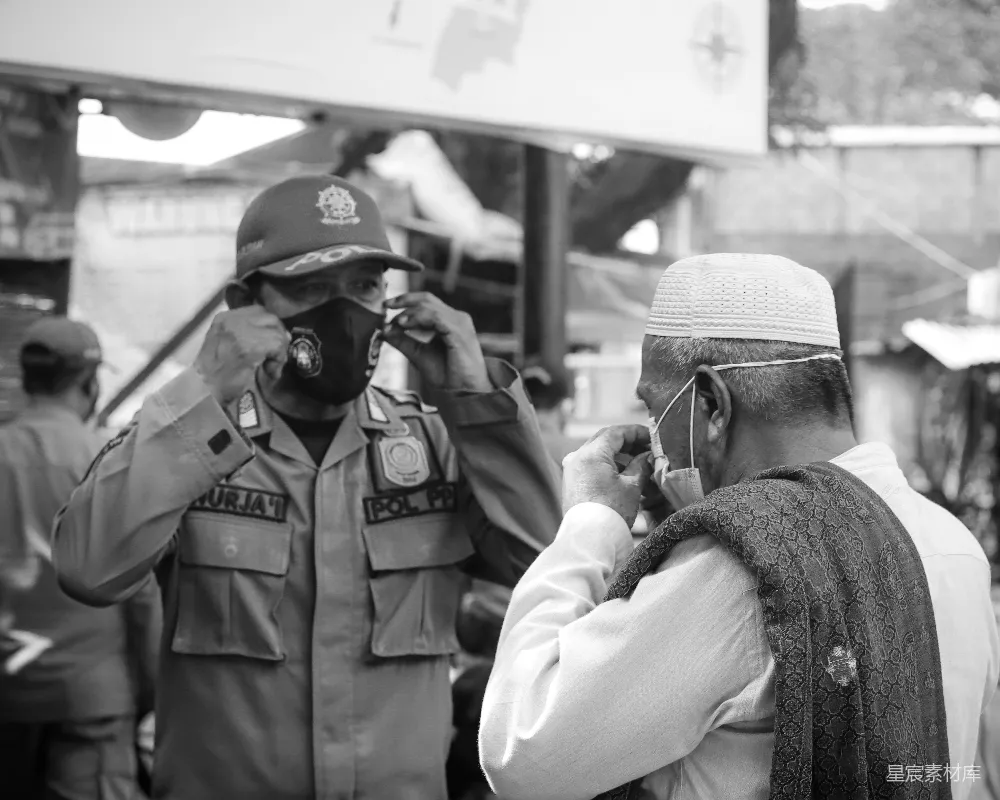 这张黑白照片展示了警察与社区居民在防疫期间互动的场景。警察正在帮助一位戴着传统头巾的老人佩戴口罩，体现了社区服务中的关怀与保护。背景模糊，突出了人物间的互动，体现了疫情期间社区成员之间的互帮互助。