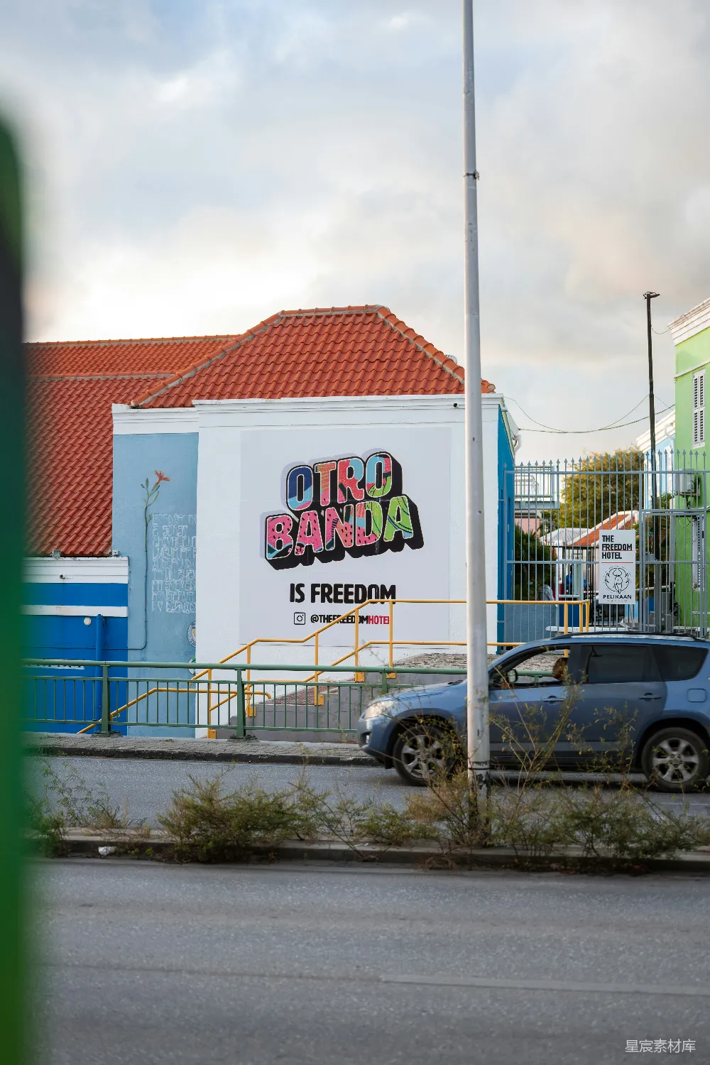 这幅色彩鲜艳的街头艺术壁画位于一座建筑的墙壁上，上面写着“OTRO BANDA IS FREEDOM”，传达了一种自由与开放的精神。壁画以蓝白为主色调，背景是城市的街景，包括红色屋顶的建筑、蓝色的栅栏和行驶的汽车，整体给人一种自由、开放和充