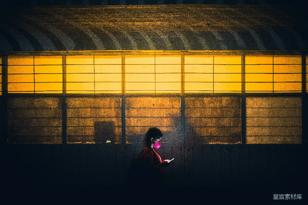 这张图片展示了一个在夜晚的城市中，一个人物的剪影，背景是温暖的灯光和建筑物的轮廓。人物手持手机，似乎在专注地查看屏幕，背景的灯光形成了强烈的光影对比，营造出一种孤独而又静谧的氛围。这个场景适合用于描述都市生活中的孤独感，或者表现夜晚城市的静
