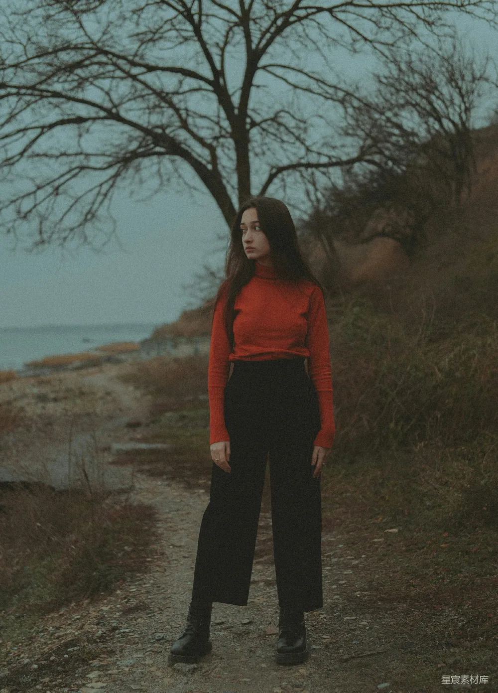 图片中，一位身穿红色高领毛衣和黑色宽腿裤的女性独自站在海边的小径上，背景是光秃秃的树木和宁静的海面。她的姿态显得有些孤寂，但又带着一种淡淡的坚定。整幅画面色调偏冷，营造出一种静谧而略带忧郁的氛围。适合用来表达孤独、思考或某种情感的表达。