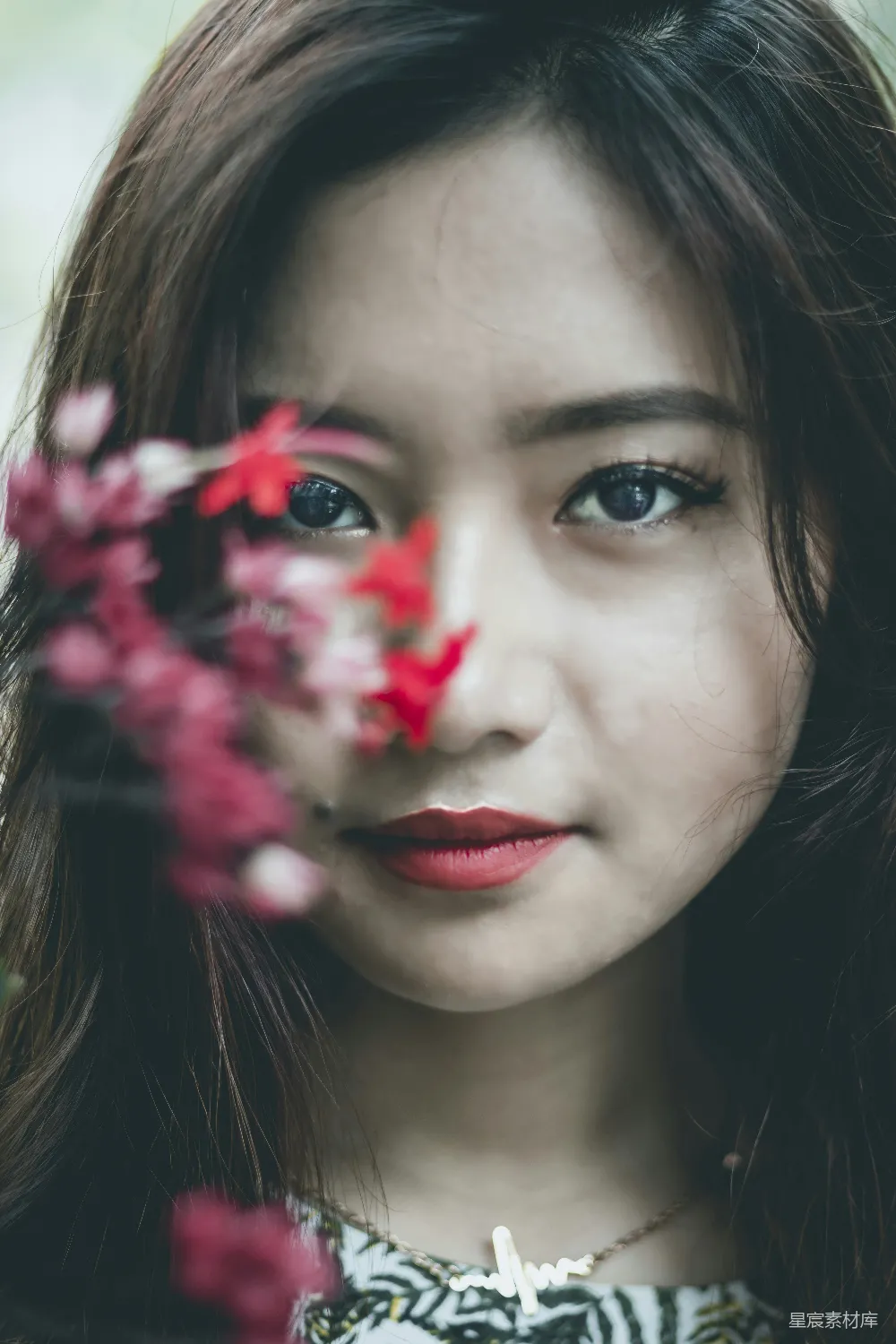 这张图片展示了一位女性，她的眼睛被一簇红色的花朵部分遮挡，营造出一种神秘而宁静的氛围。她的妆容精致，红色的唇色与花朵相得益彰，背景模糊，突出了人物的面部特征。这种风格的图片适合用于展示自然美、情感表达以及日常生活中的美好瞬间。
