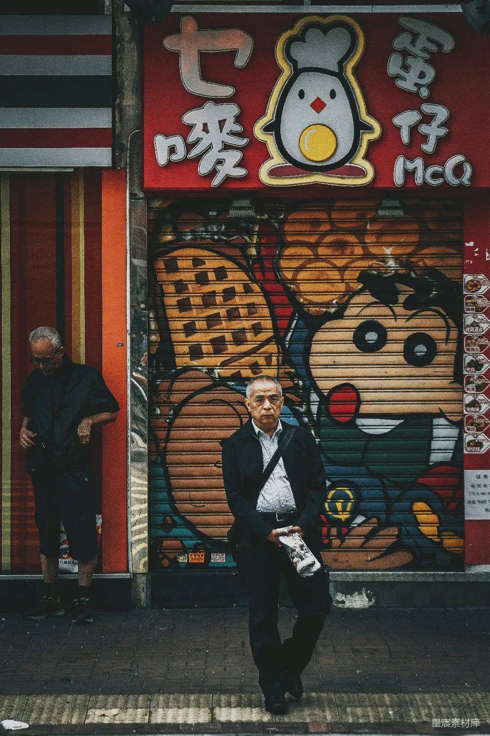 这张图片展示了一位老男人坐在一个色彩斑斓的涂鸦墙前，背景是红色的招牌和卡通鸡的图案。他的姿态和周围环境形成了对比，给人一种静谧而深思的感觉。这种场景常见于城市中的街角巷尾，是城市生活和街头艺术的生动体现。