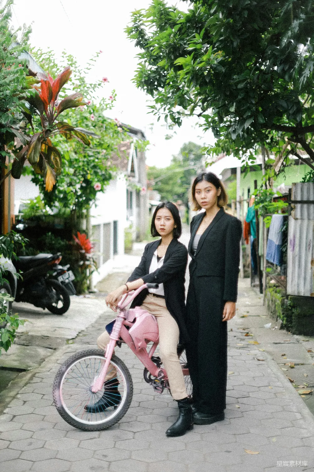 这张图片展示了两个姐妹站在城市街道上，其中一位坐在粉色的自行车上，另一位则站在一旁。背景是充满绿植和房屋的街道，充满了自然与城市生活的交织。姐妹们的服饰简约而时尚，营造出一种轻松而美好的氛围。适合用来展示友情、城市生活或旅行的主题。