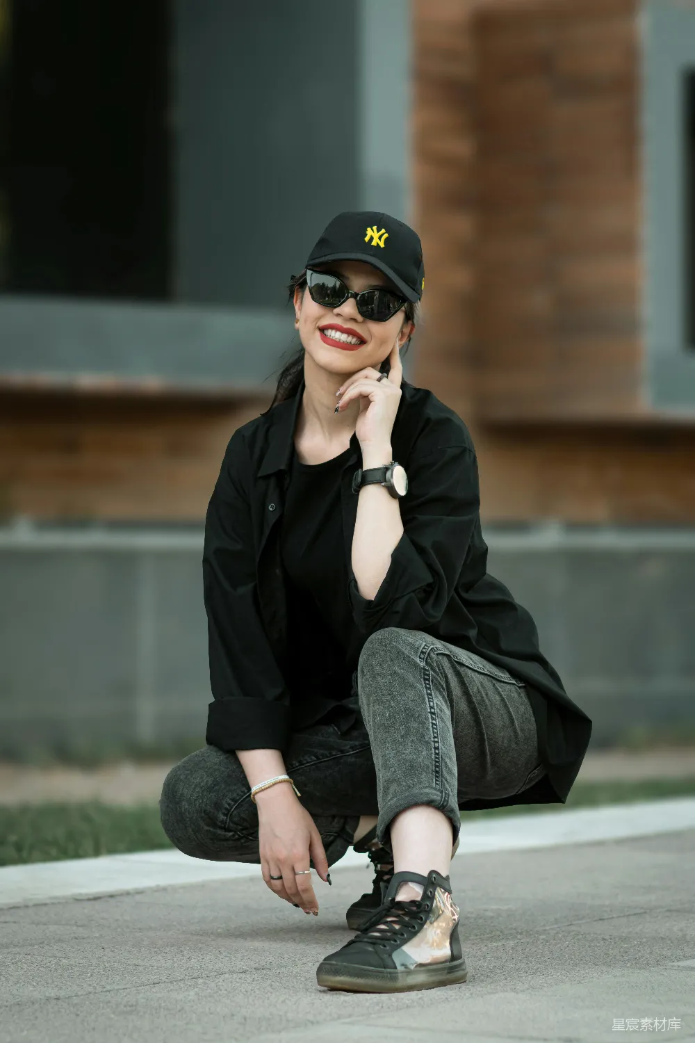 图片展示了一位女性在户外环境中，身穿黑色T恤、外套和牛仔裤，搭配黑色棒球帽、太阳镜和独特设计的运动鞋。她以蹲坐的姿势，双手轻触脸部，展现出自信和优雅的气质。背景是模糊的建筑物，突显了人物的时尚感和现代都市氛围。适合用于时尚、街头风、休闲等场