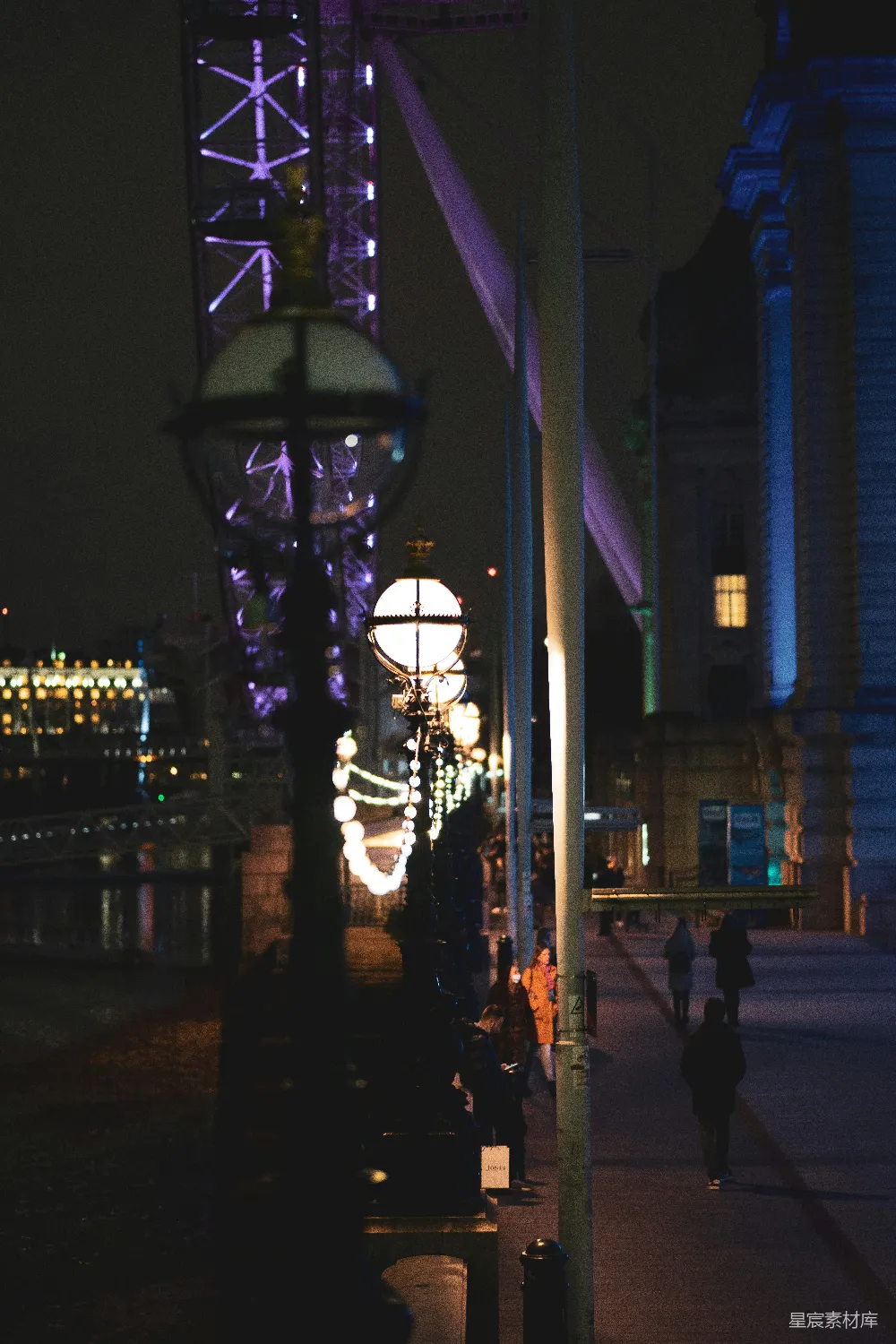 这张图片展示了伦敦夜景下的街灯，背景是著名的伦敦眼，灯光与建筑在夜色中显得格外迷人。街道上行人稀疏，营造出一种宁静而优雅的氛围。适合用来展示城市夜景和旅游景点。