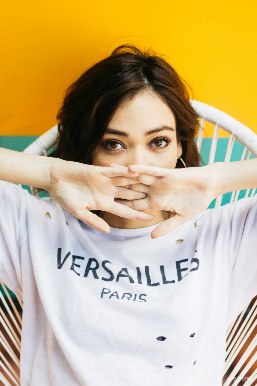 图片展示了一位穿着带有'VERSAILLES PARIS'字样的破洞T恤的女性，她用双手遮住面部，形成了一个有趣的创意手势。背景是鲜艳的黄色和绿色，营造出一种活泼而时尚的氛围。适合用于时尚、创意或生活类的素材展示。