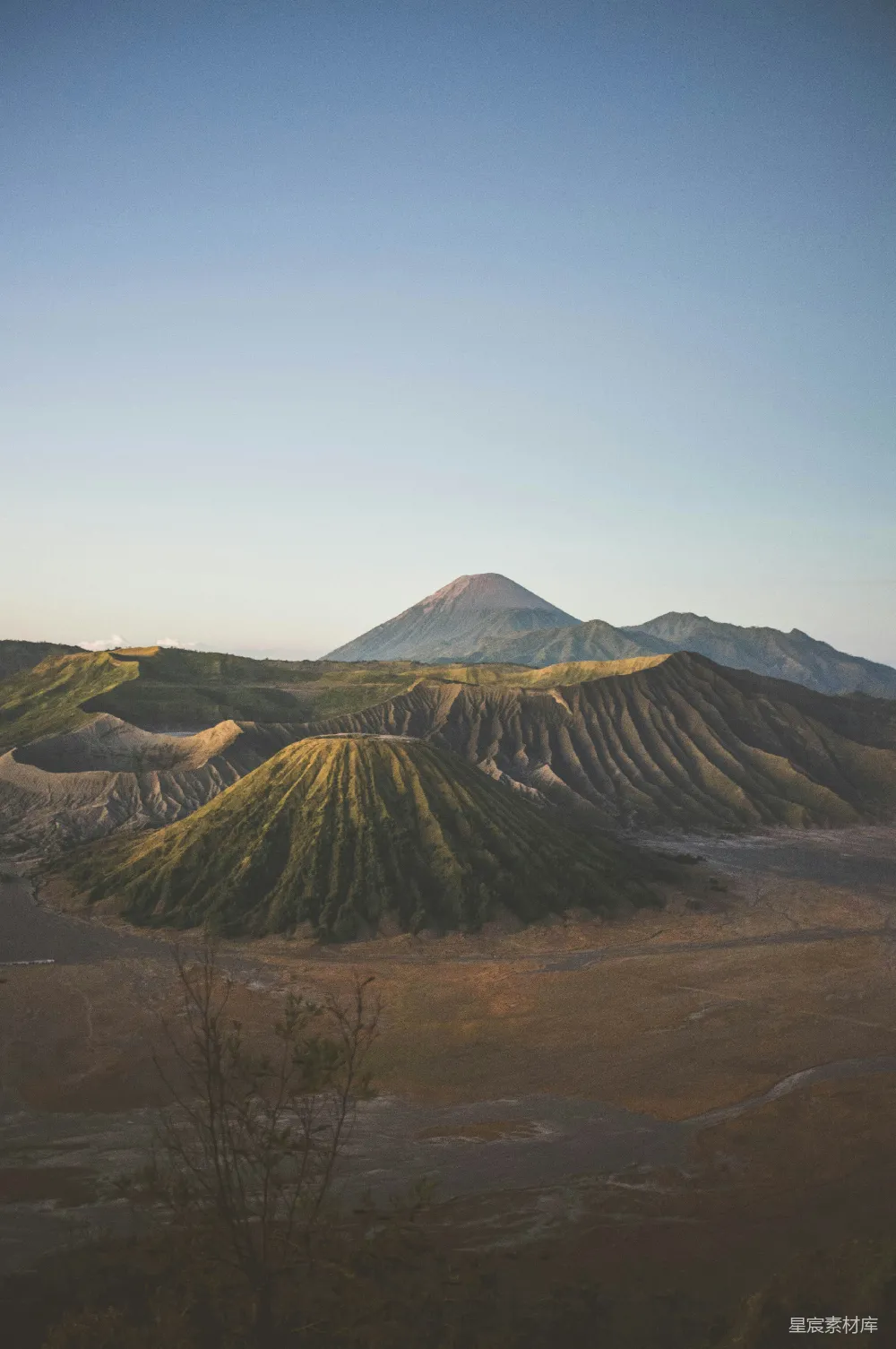 这张图片展示了火山和山丘的壮丽景色，背景是蓝天，火山的轮廓清晰可见，前景是起伏的山丘，植被丰富。整体画面色彩柔和，给人一种宁静而神秘的感觉，适合用作户外探险、自然风光和旅行宣传的背景图。