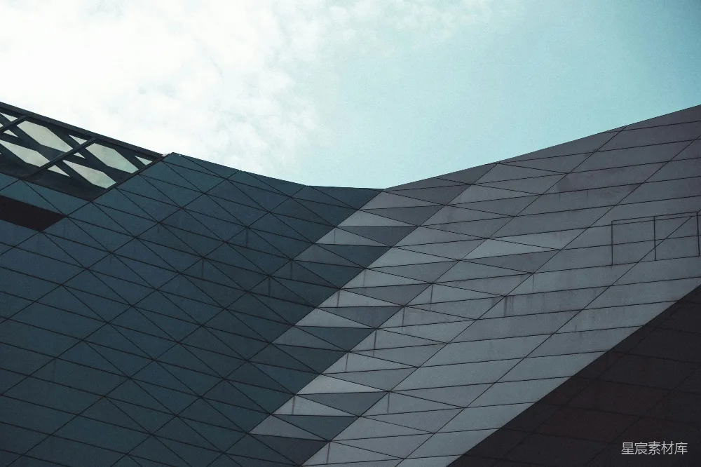 这张图片展示了现代建筑的几何结构，建筑表面由三角形图案构成，形成一种简洁而富有节奏感的视觉效果。背景是蓝天白云，使得建筑显得更加突出。这种风格的建筑常见于城市公共建筑或商业中心，适合用于展示或装饰。