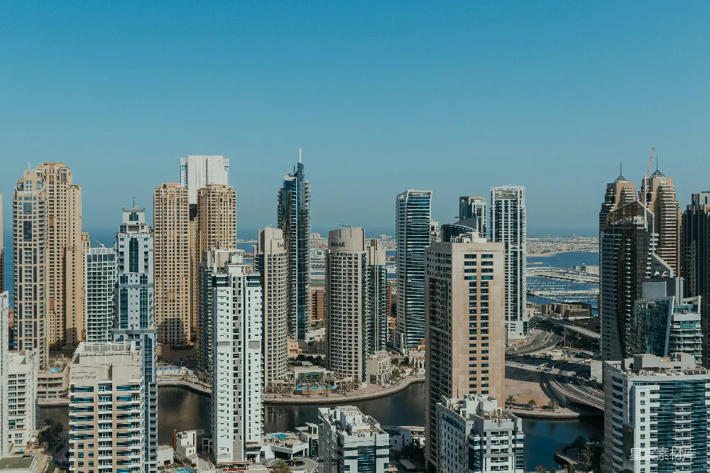 这张图片展示了迪拜的摩天大楼群，从高空俯瞰视角可以清晰地看到密集的高层建筑，背景是蓝天和远处的地平线。这些建筑风格多样，既有现代感十足的玻璃幕墙，也有传统色调的建筑。图片中还有一条水道穿插其中，增加了画面的层次感。整体风格现代而充满活力，适