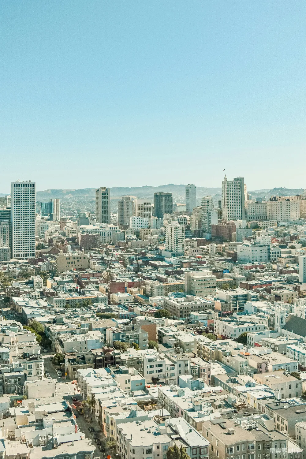 这张图片展示了阳光明媚的城市全景，天空湛蓝，建筑物错落有致，屋顶在阳光下泛着柔和的光芒。城市中既有现代化的高楼大厦，也有低矮的住宅区，远处可以看到连绵的山脉。整体画面给人一种宁静而美丽的视觉享受。