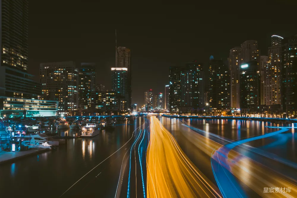 这张图片展示了夜晚的城市景观，高楼大厦灯火辉煌，街道上的车流形成光的轨迹，倒映在水面上，形成美丽的光绘效果。适合用作都市生活的背景图片，或用于夜间城市风光的展示。