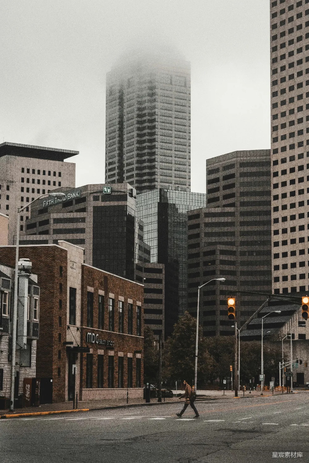 图片展示了一座城市的街景，高楼大厦在雾气中若隐若现，街道上行人匆匆走过，给人一种繁忙与宁静交织的感觉。这幅图像通过冷色调和朦胧的雾气，营造出一种神秘而现代的都市氛围。适合用于城市生活、建筑设计或自然与城市融合的主题展示。