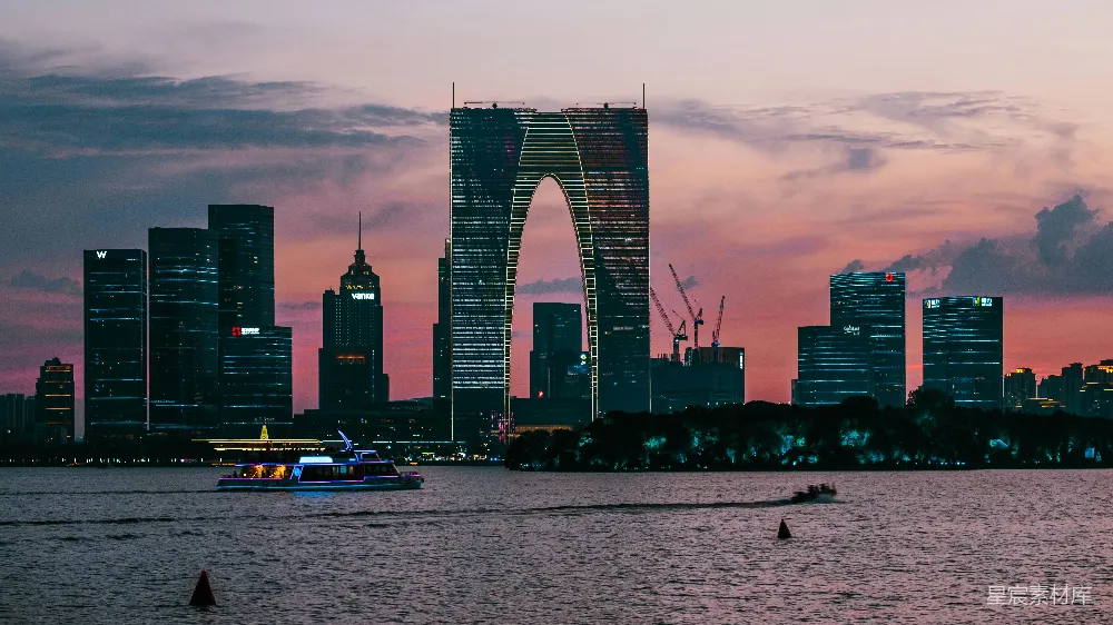 这张图片展示了日落时分的城市天际线，高楼大厦在夕阳的映衬下显得格外壮观。天空呈现出柔和的橙色和粉色调，与城市的灯光交相辉映，形成了一幅美丽的城市风景画。前景中有几艘船在河面上行驶，增添了画面的动感。