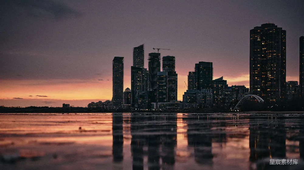 这张图片展示了日落时分的城市天际线，高楼大厦在夕阳的余晖中剪影，倒映在平静的水面上，营造出一种宁静而美丽的都市夜景。