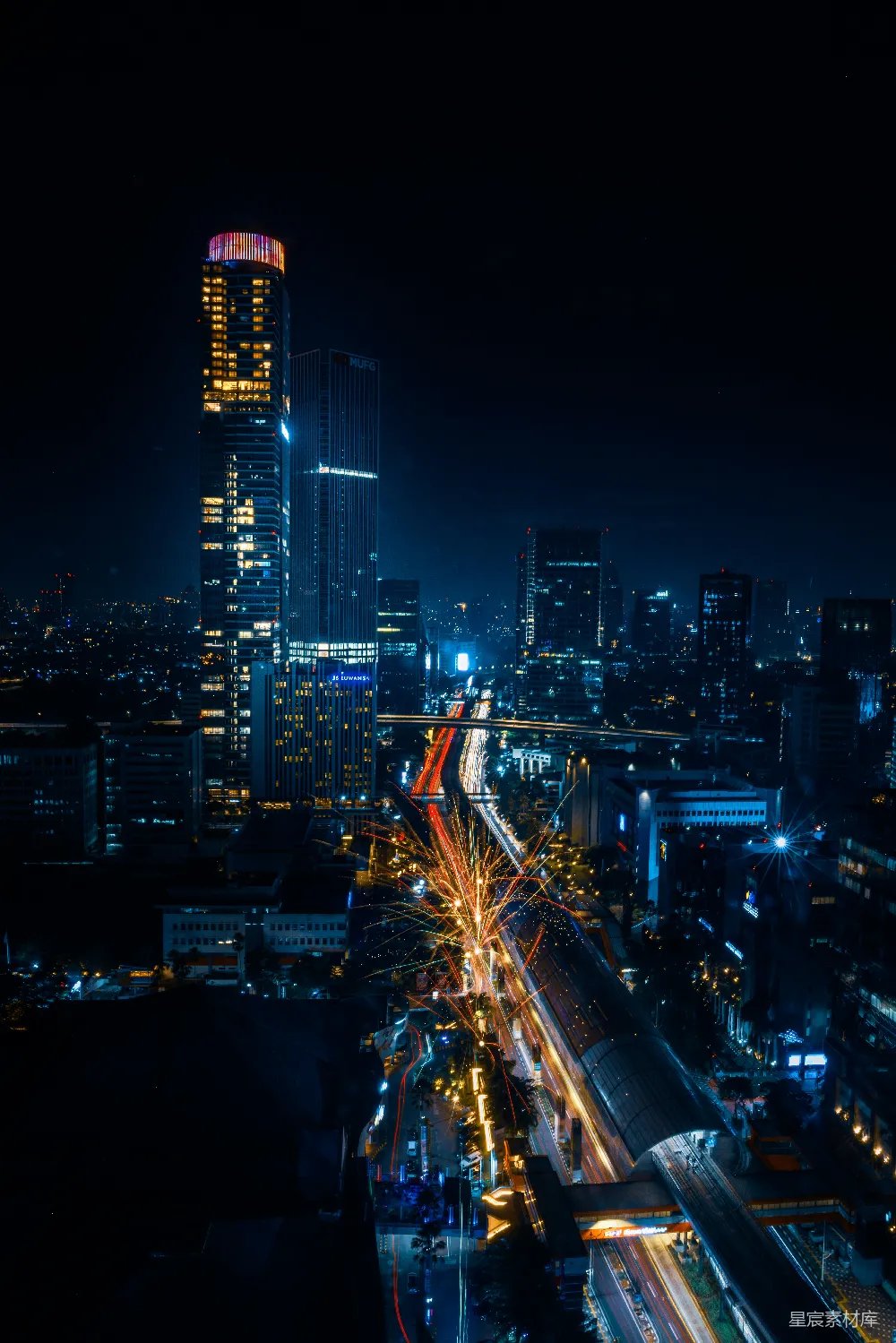 这张图片展示了一个城市的夜景，高楼大厦灯火通明，道路上的车流形成光带，天空中绽放着五彩斑斓的烟花。整个画面充满了现代都市的繁华与活力，适合用作城市风光的宣传图片。