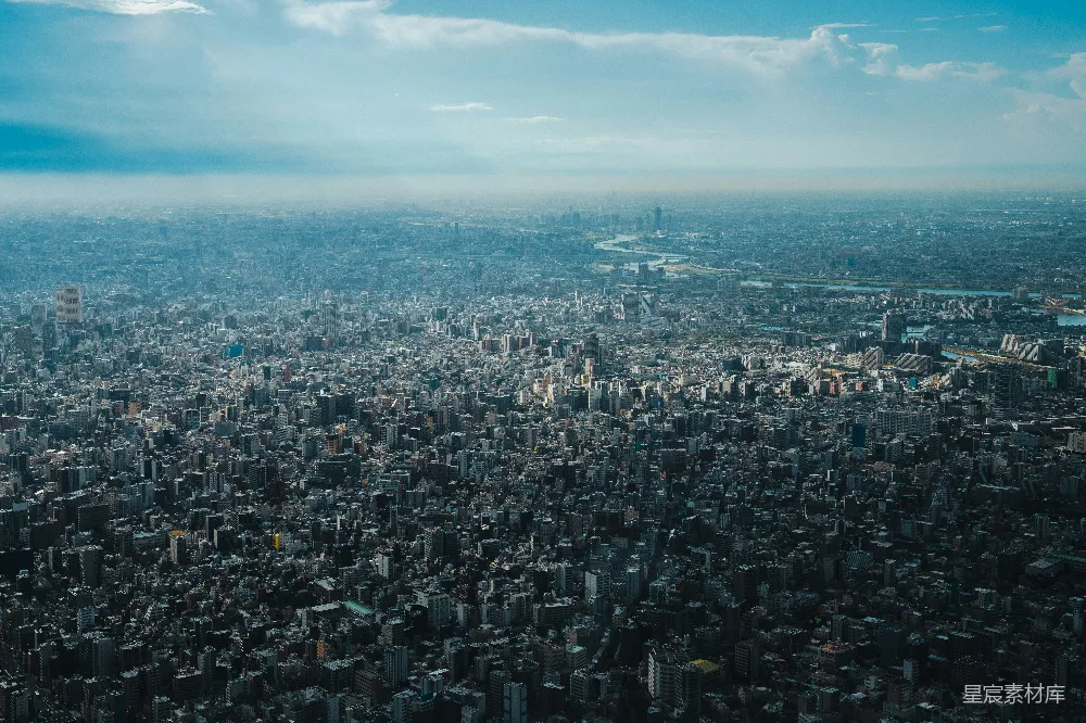这张图片展示了东京的俯瞰全景，天空中散布着几朵白云，城市中密布着高楼大厦，远处可以看到河流蜿蜒穿过。图片展示了东京繁华的城市风貌，适合用于旅游、城市规划和地理教育场景。
