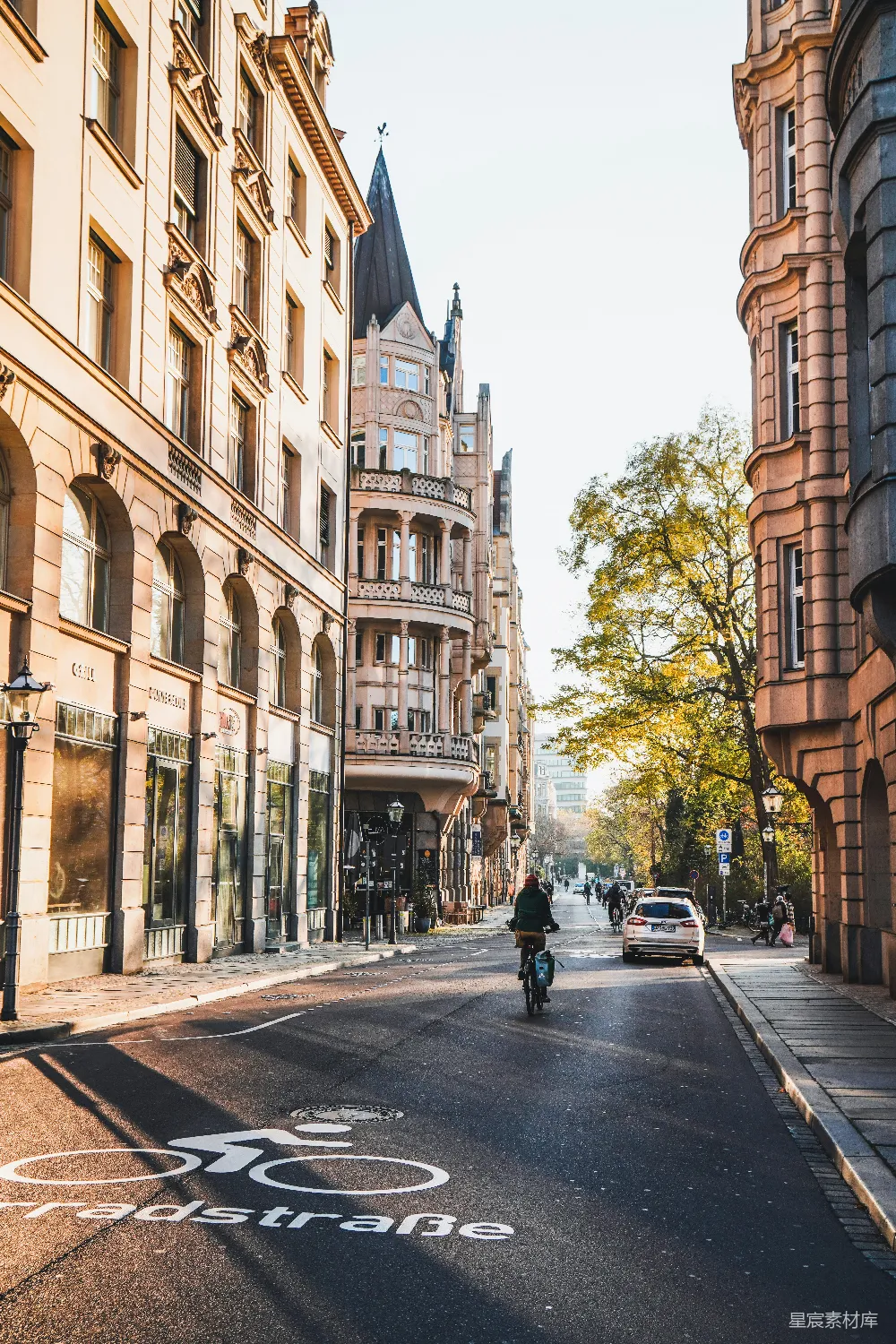 图片展示了一条位于德国古城的街道，两旁是历史悠久的建筑。街道中间有一条自行车道标志，道路上有行人和骑车人，远处可以看到一座尖顶教堂。树木郁郁葱葱，阳光洒在街道上，营造出一种宁静而美好的氛围。适合用于展示欧洲古城的日常生活场景。