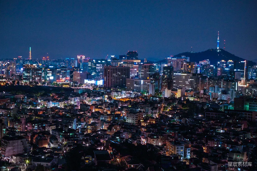 这张图片展示了夜晚的城市繁华景象，高楼大厦灯火辉煌，错落有致，远处的山丘上点缀着几盏灯光，增添了画面的层次感。天空深邃，城市的灯光与夜空形成鲜明对比，展现了都市夜景的迷人魅力。适合用于城市风光、夜间摄影和现代都市主题的背景图。