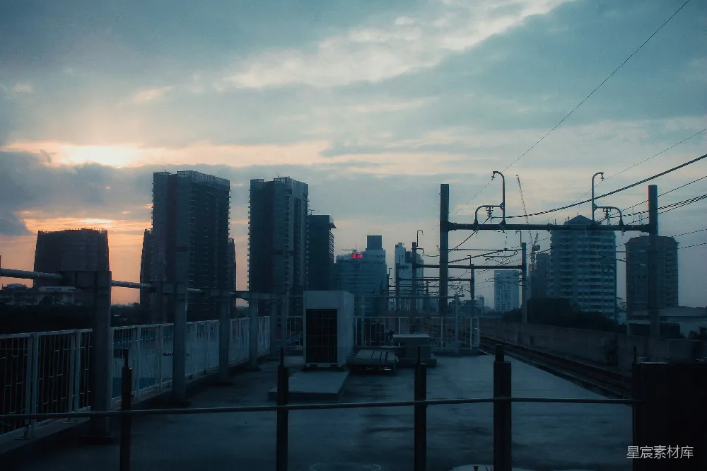 这张图片展示了一个城市在日落时分的景象，天空被橙色的余晖染成温暖的色调。远处的高楼大厦在夕阳的映衬下显得格外醒目，而前景中的铁路轨道和电线则增添了一丝工业的韵味。整体色调偏冷，给人一种宁静而深思的感觉。