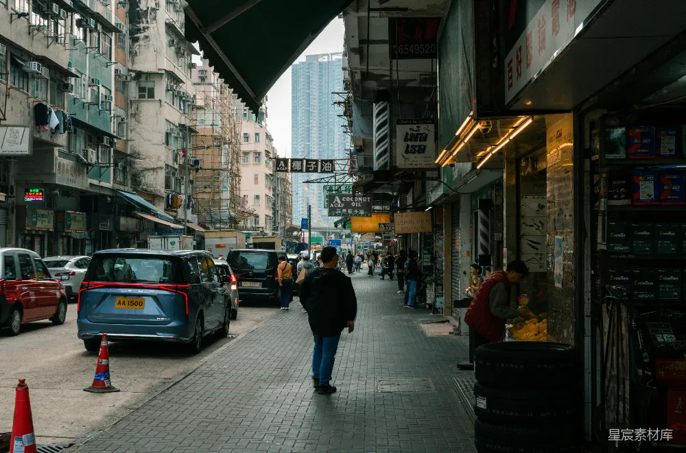 这张图片展示了一条香港的典型街道，两侧是密密麻麻的商店和居民楼。街道上行人和车辆穿梭，氛围繁忙而充满生活气息。建筑物上的招牌和广告牌清晰可见，街道两旁堆放着各种商品，给人一种紧凑而充满活力的感觉。