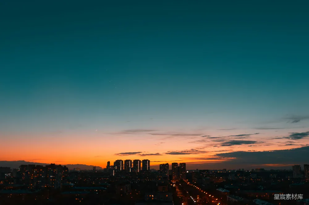 这张图片展示了城市在夜幕降临时的宁静景象。天空由深蓝渐变至橙色，城市中的高楼大厦在灯光的映衬下形成剪影，远处的山脉在夕阳的余晖下显得格外神秘。整个场景给人一种静谧而深邃的感觉，适合用于城市生活、自然风光或摄影教程的背景素材。
