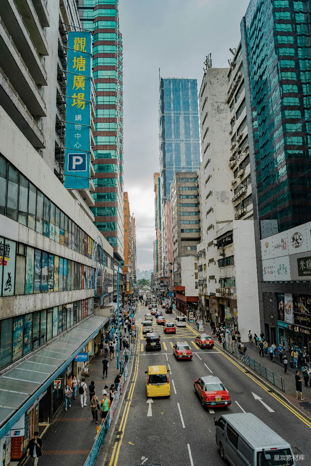 这张图片展示了一条繁忙的都市街道，两旁是高楼大厦和商铺，街道上车流不息，行人穿梭其间。建筑物的外观多样，既有现代化的玻璃幕墙，也有复古的工业风格。天空阴沉，给人一种忙碌而略显压抑的感觉。适合用于描述都市生活、商业区或现代城市风貌的主题。