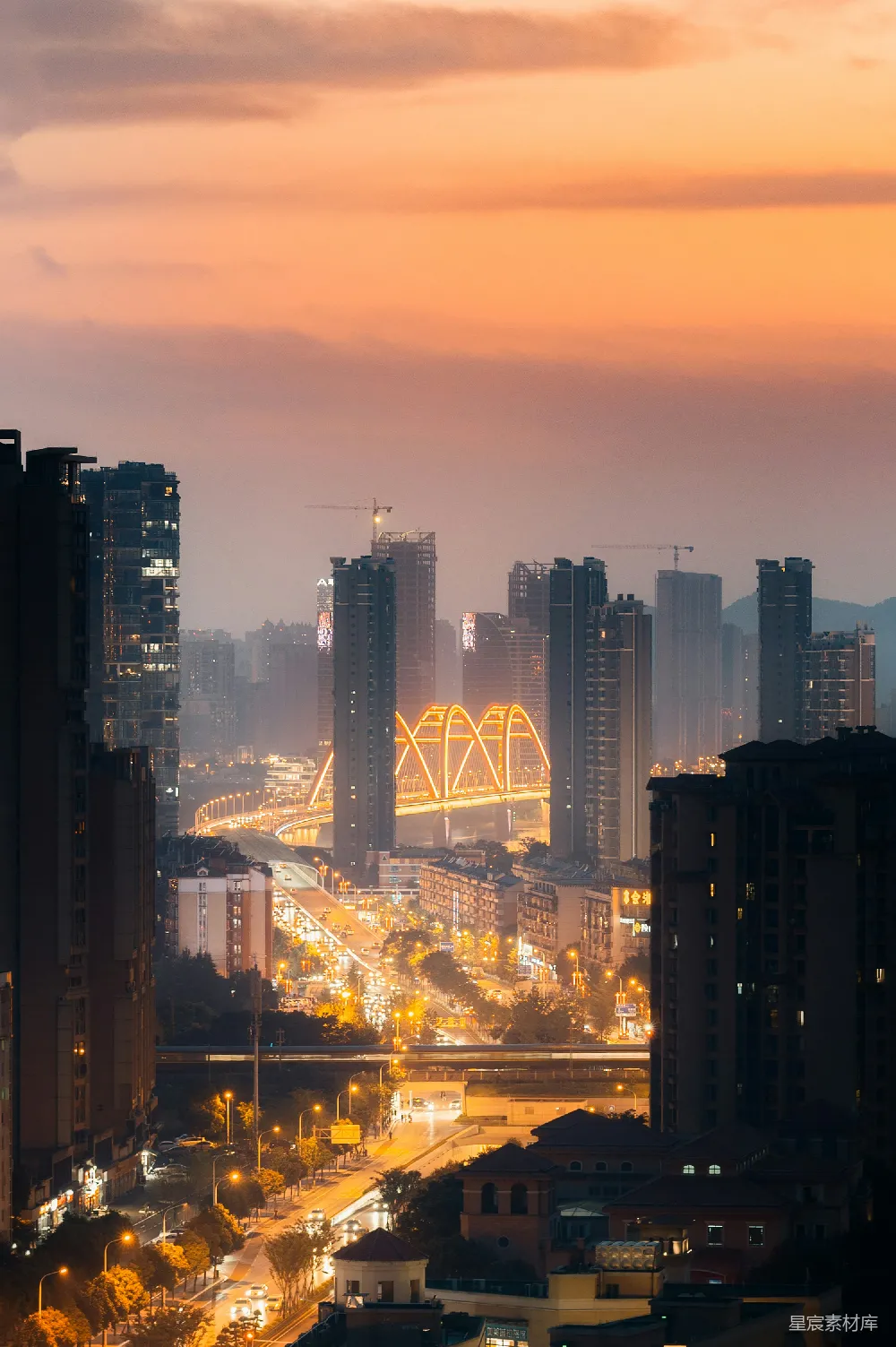 这张图片展示了日落时分的城市天际线，夕阳的余晖洒满天空，为高楼大厦披上了一层金黄色的光辉。城市中的桥梁灯火通明，与远处的山峦形成鲜明对比，营造出一种宁静而美丽的都市夜景。