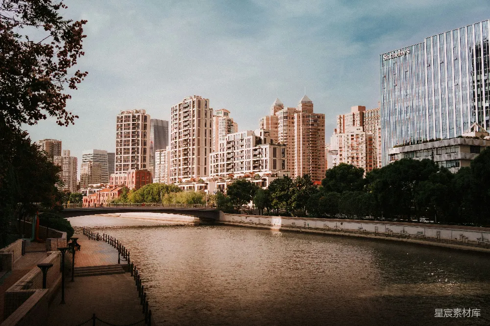 这张图片展示了一条河流穿过城市的景象，河岸边有树木和步道，背景是高楼大厦。天空晴朗，光线柔和，营造出一种宁静而美好的氛围。适合用于城市生活、休闲旅游或都市风光的展示。