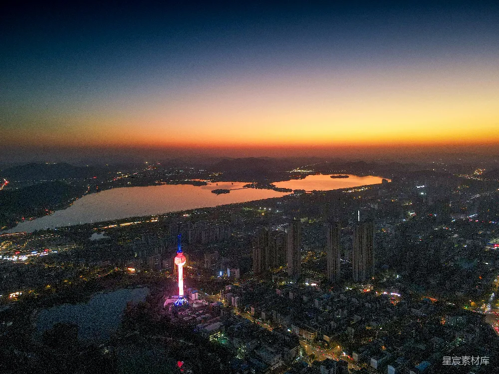 这张图片展示了一个城市的夜景，湖水在夕阳的余晖中泛着金色的光，湖畔的城市建筑群灯火辉煌，形成了一幅美丽的城市夜景图。画面中央有一座独特设计的塔楼，其灯光与周围的夜景相得益彰，给人一种现代都市的科技感与活力。这种场景适合用于城市旅游宣传、夜间