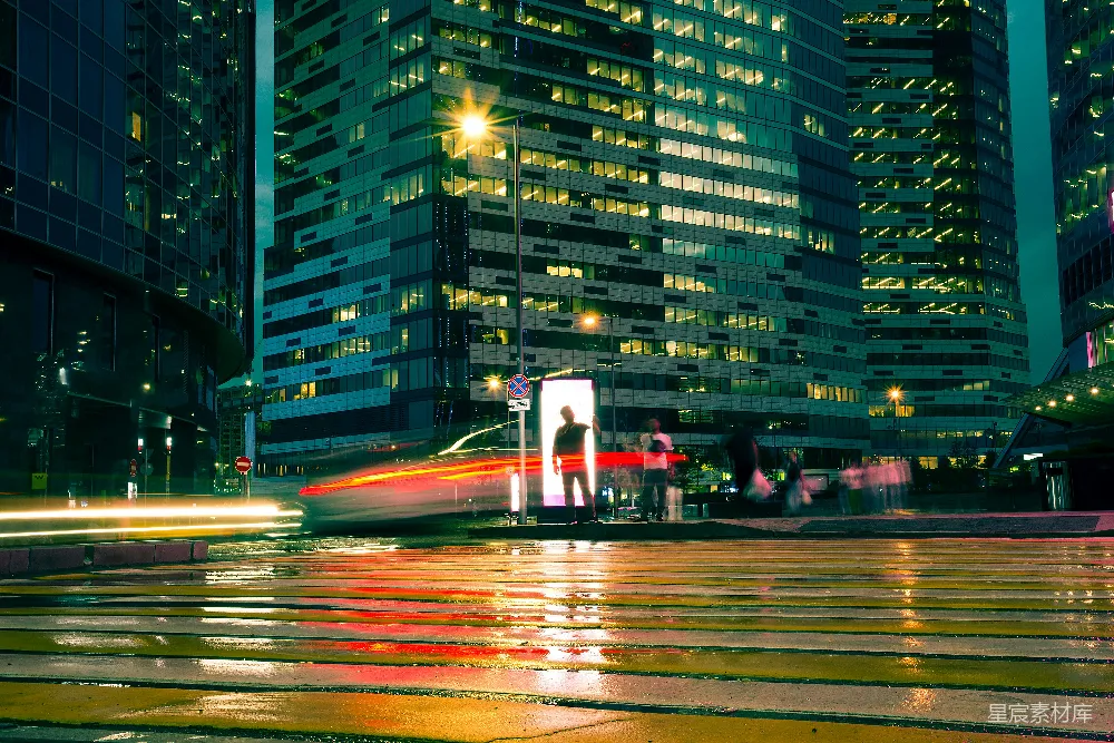 这张图片展示了夜晚城市中繁忙的街景。雨水打湿了地面，反射出车辆和街道的光亮。高楼大厦的窗户透出温暖的灯光，形成一片片光斑。前景中，行人和车辆在光与影的交织中穿梭，构成一幅充满动感与现代感的都市夜景。