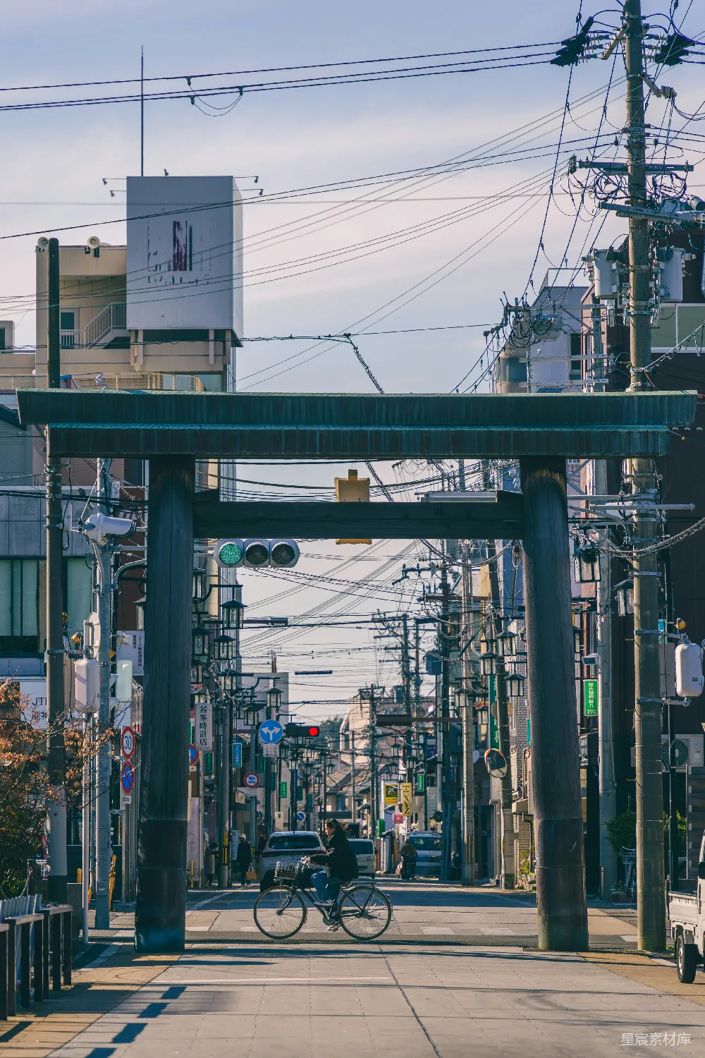 这张图片展示了一座传统的神社鸟居，被现代城市的电线和建筑物环绕。一名骑自行车的人穿过鸟居，增添了一丝生活气息。背景中的建筑和电线展示了都市的复杂性和多样性，而鸟居则象征着传统与现代的融合。适合用于展现城市文化、传统与现代交织的主题。