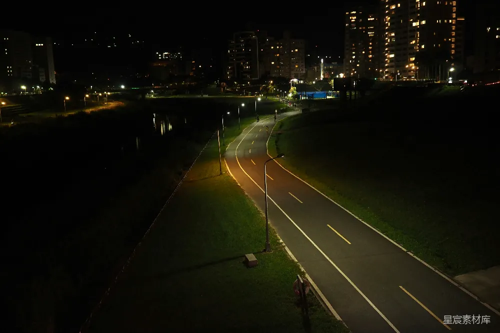 这张图片展示了一条城市中的道路在夜晚的景象。道路两旁是绿植和路灯，远处可以看到灯火通明的建筑物，营造出一种宁静而充满活力的氛围。