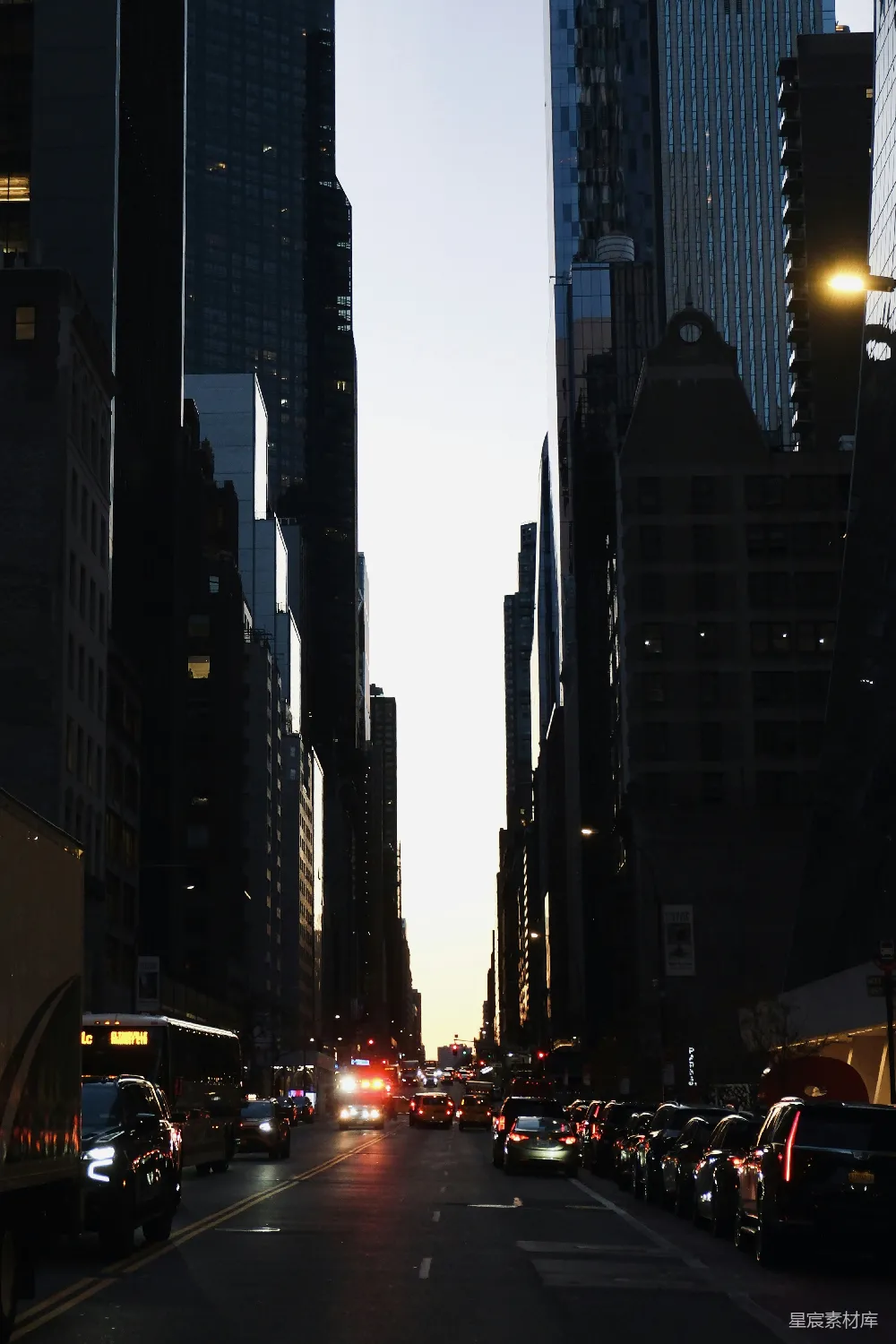 这张图片展示了一个城市的街道，在夕阳的余晖中，高楼大厦的剪影显得格外突出。街道两旁停满了车辆，远处的车辆灯光点亮了夜幕的边缘。这幅画面捕捉了都市生活的宁静与繁华之间的对比，给人一种时间静止的感觉。