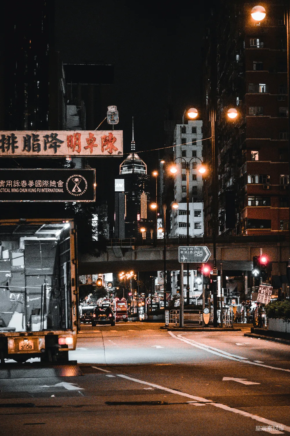 这张图片展示了香港夜晚的街道景象，灯光柔和，招牌上印有中文，远处是现代化的高楼大厦。街道两旁是各种建筑，交通灯和路灯照亮了道路，给人一种静谧又充满活力的感觉。适合用于城市夜景、香港文化和街道风景等主题。