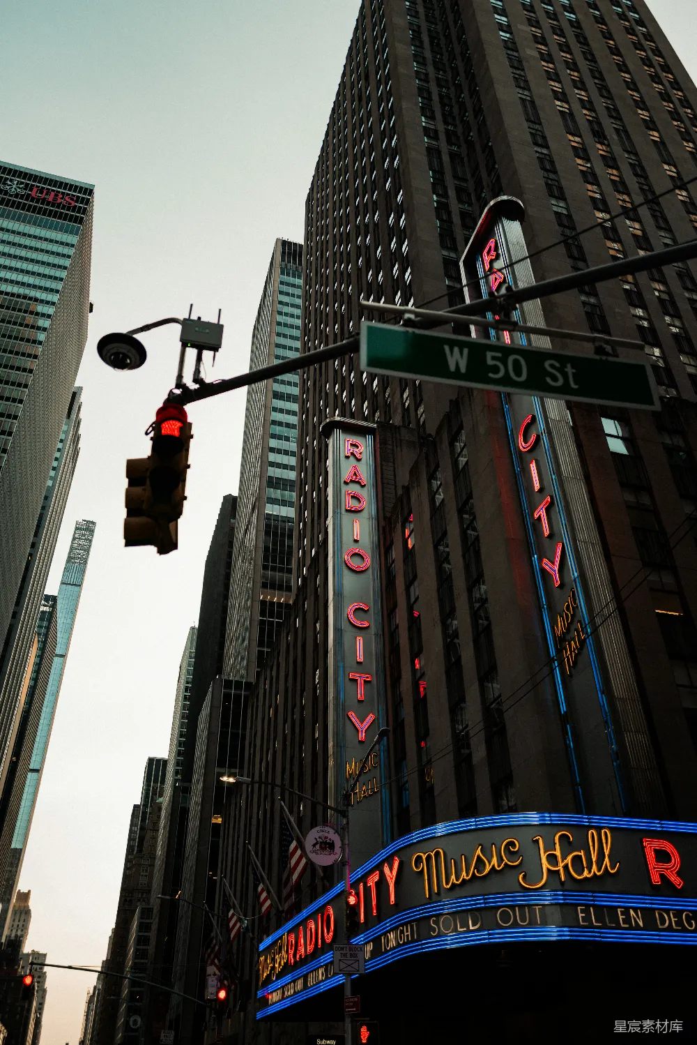 这张图片展示了纽约第五大道上的标志性建筑——Radio City Music Hall。夜晚的霓虹灯光与现代摩天大楼形成鲜明对比，营造出一种动感的都市氛围。街灯和交通信号灯的红色与建筑上的蓝色霓虹灯形成视觉上的冲突，进一步增强了画面的视觉冲