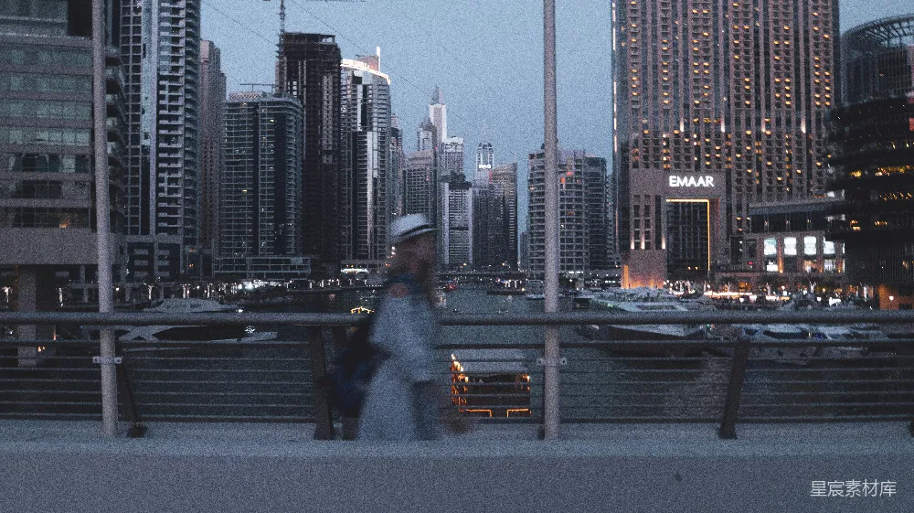 这张图片展示了一位行人在城市夜景中的剪影，背景是灯火辉煌的摩天大楼和繁忙的港口。画面色调偏冷，给人一种宁静而又充满活力的感觉，适合用于现代都市生活的主题。